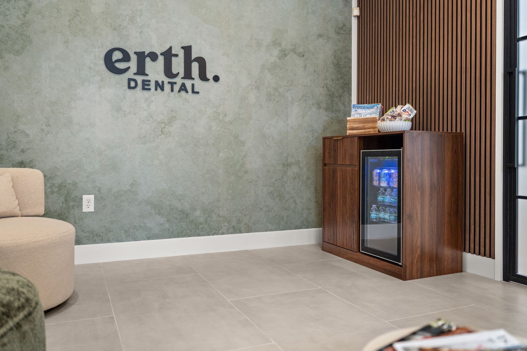 A walnut refreshment cabinet with a drinks fridge, snacks and magazines beside the erth. dental sign