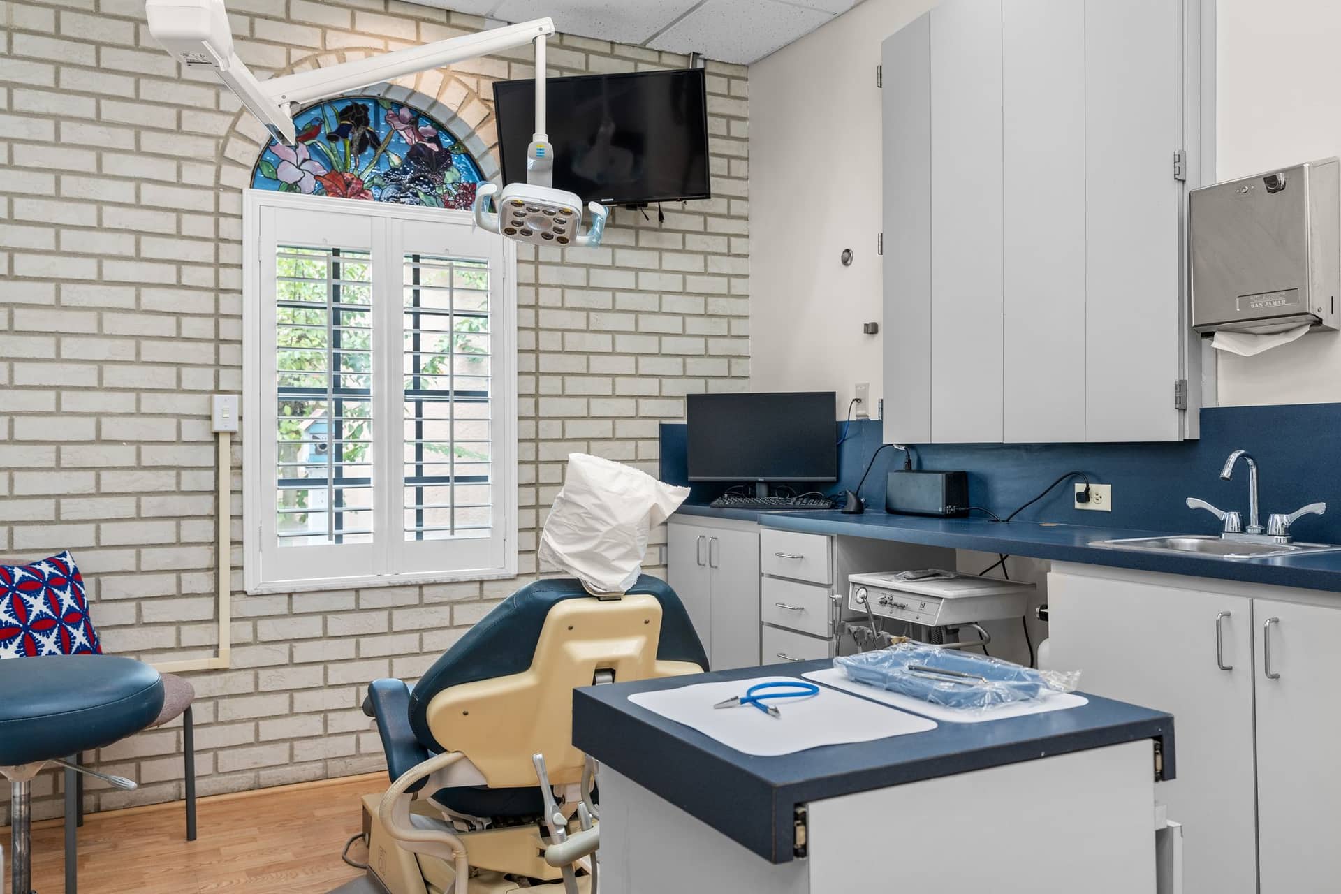 A treatment room at erth. dental with a dental chair, a stained glass arch above a shuttered window and blue countertops